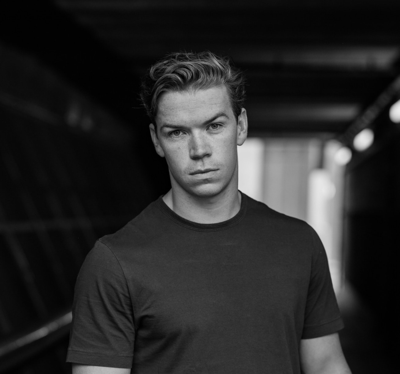 Siyah beyaz portrede koyu tişörtlü Will Poulter, loş bir koridorda kameraya bakıyor.
