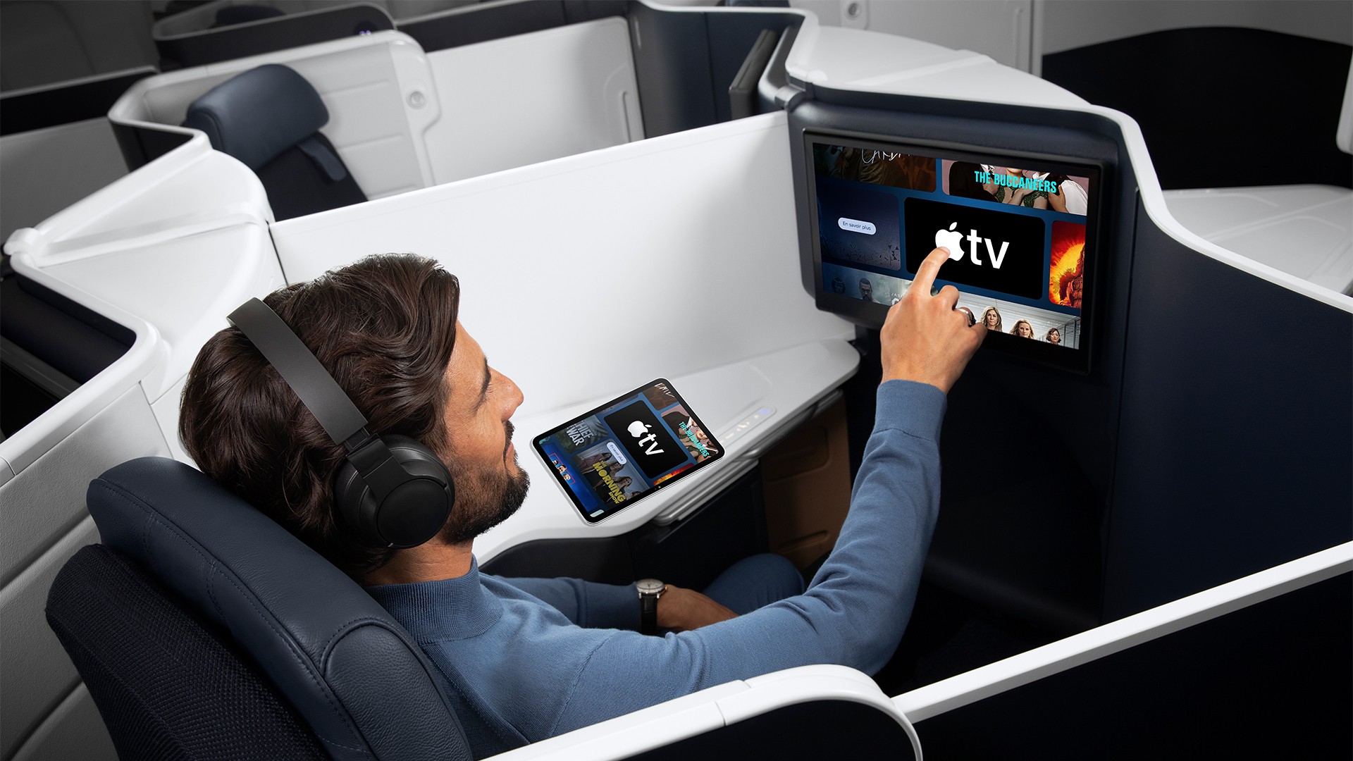 Uçakta iş sınıfı koltuğunda oturan bir yolcu, önündeki ekranda Apple TV uygulamasına dokunuyor.