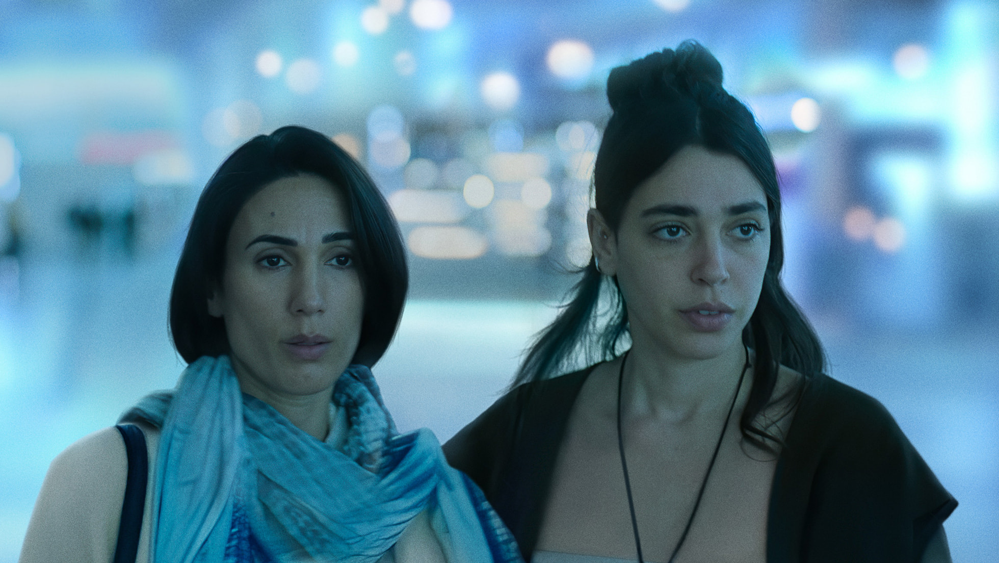 Dizi karesinde bir havalimanı terminalinde yan yana duran, endişeli iki kadın.