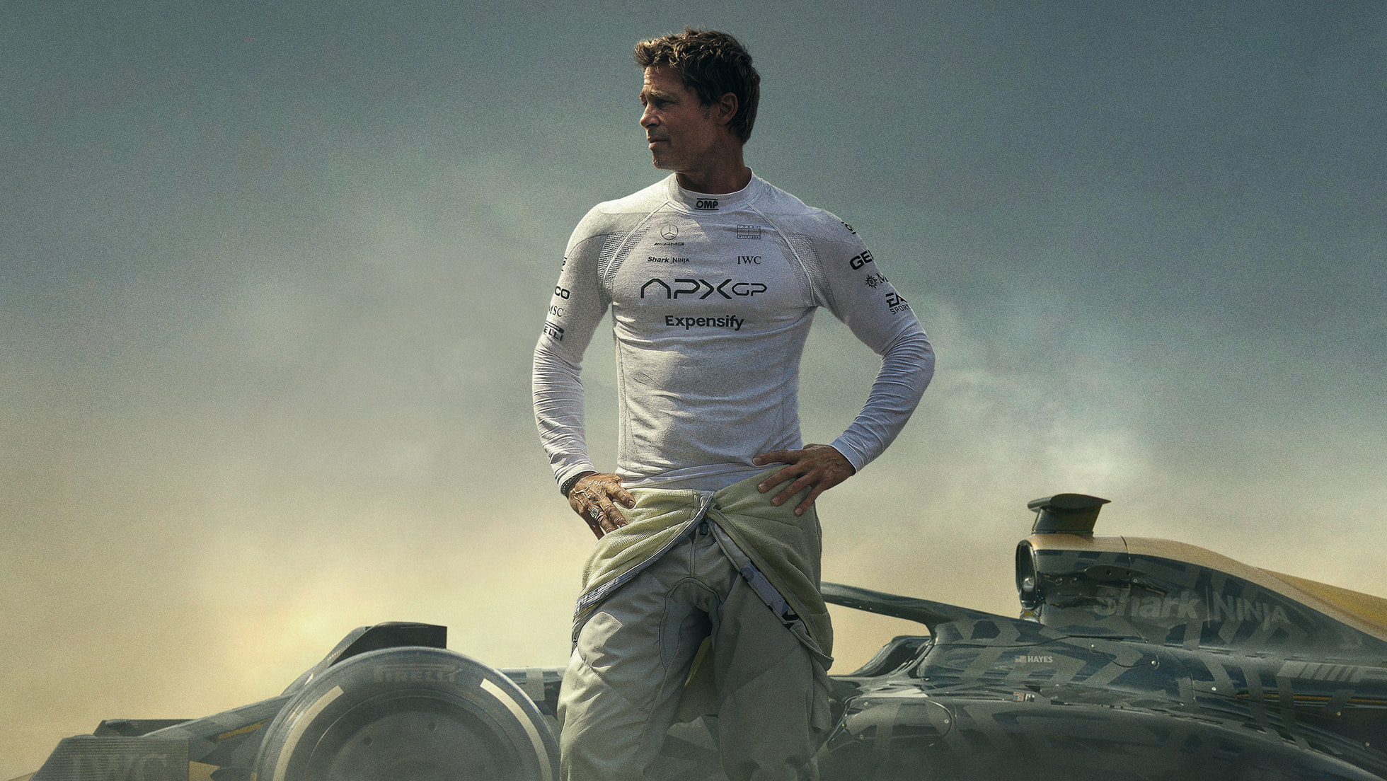 F1 filminden bir kare: yarış tulumunu beline indirmiş bir sürücü, arkasında Formula 1 aracı duruyor.