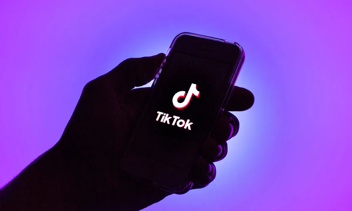 Karanlıkta bir el, ekranında TikTok logosu parlayan bir telefonu tutuyor; arka plan mor.