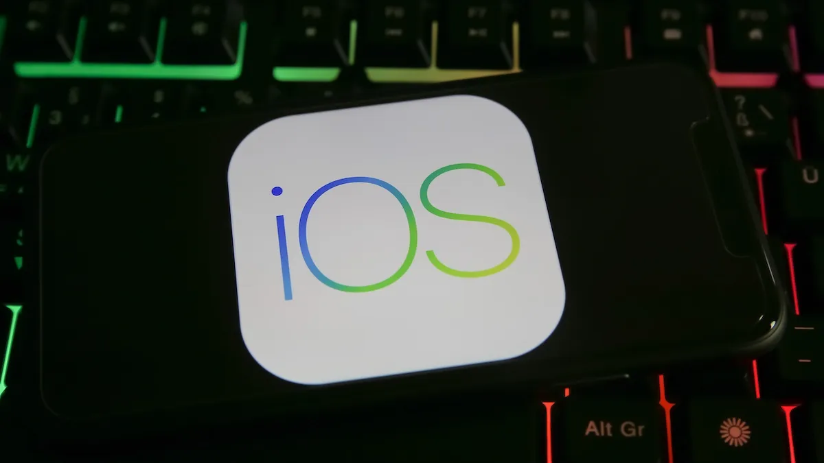 Renkli ışıklı bir klavyenin üzerinde duran, ekranında iOS simgesi görünen bir telefon.